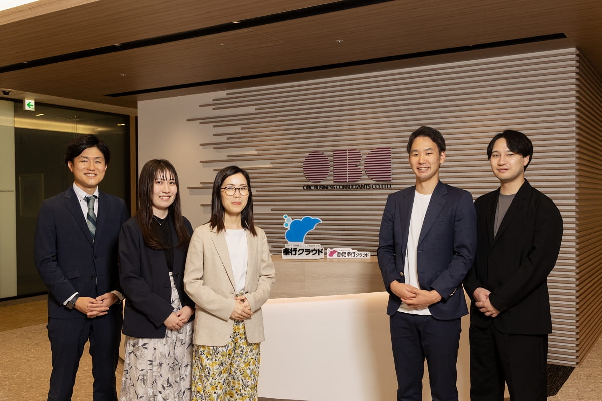 OBCのロゴの前に立つOBC 本澤浩樹さん、山口まゆ子さん、坂本麻美さんと、CINC コンサルタントの藤原清誠、小櫃裕也の写真。